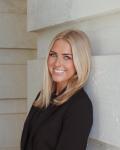 A young woman with blond hair wearing a black suit and leaning against a wall