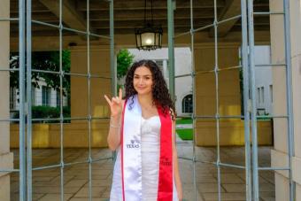 Chloe Landau Graduation Picture, white dress with red stole, standing in front of iron doors, with hook 'em hand gesture